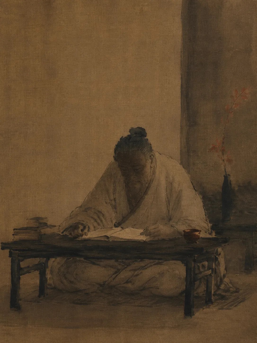 An ink painting on aged silk of an elderly scholar in plain undyed robes, seated cross-legged at a low table, head lowered over an open book. A branch stands in a dark vase behind him.